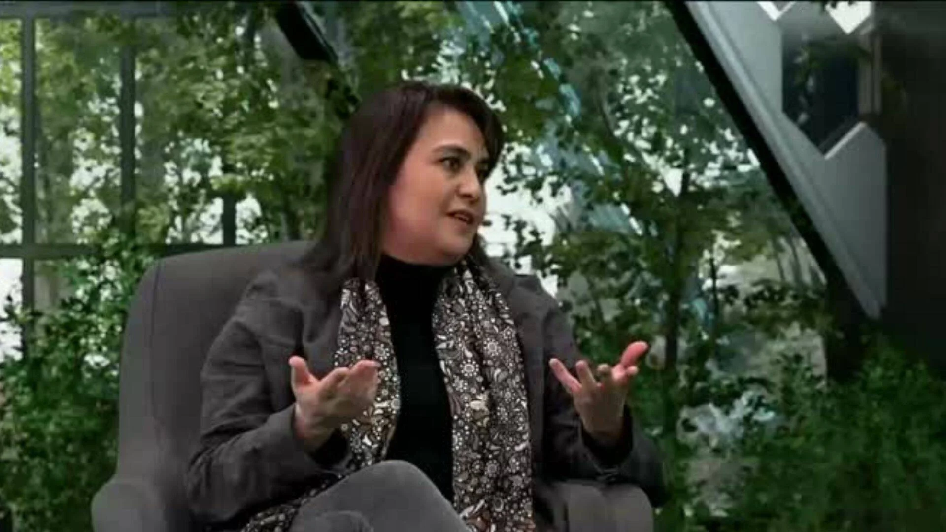 Mujer con blazer gris y pañuelo floral gesticula mientras habla sentada en un estudio de televisión con vegetación de fondo.