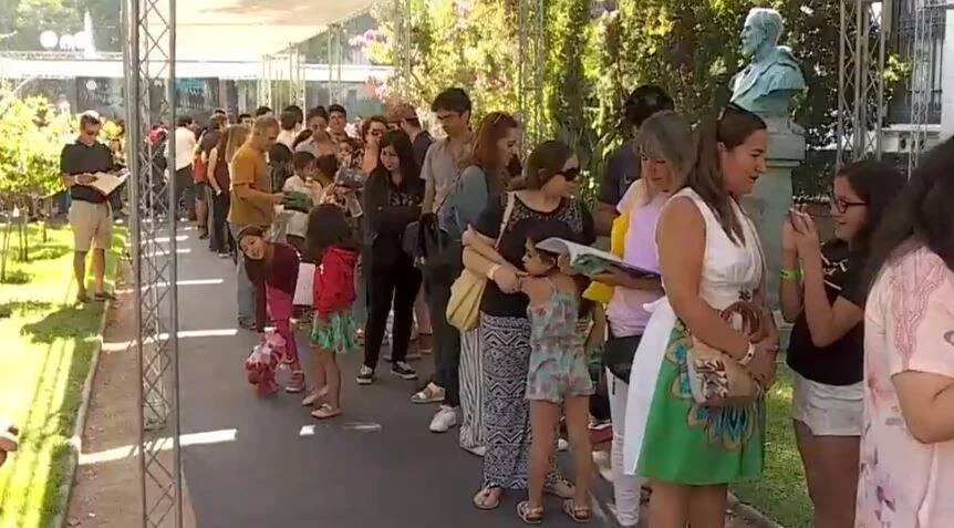 Numerosas personas con libros en mano hacen fila en un paseo arbolado durante una feria del libro al aire libre.