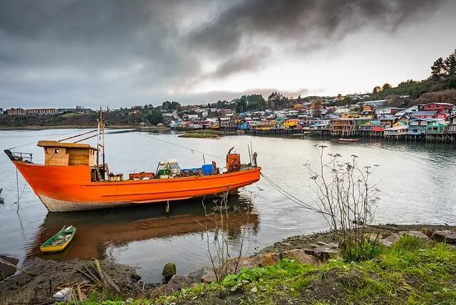 Embarcación de madera color naranja varada en la orilla con casas sobre pilotes al fondo bajo cielo nublado.