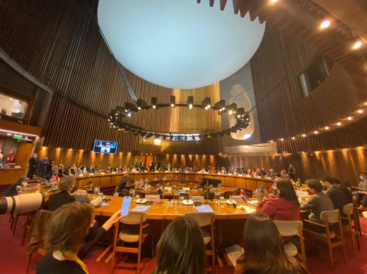 Delegados de múltiples países sentados en mesa redonda durante una reunión multilateral en un salón de conferencias con banderas nacionales.