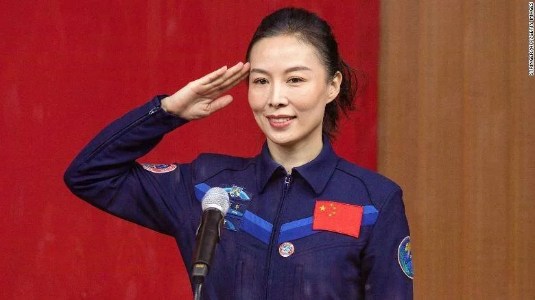Astronauta con traje espacial azul y bandera de China hace el saludo militar frente a un micrófono.