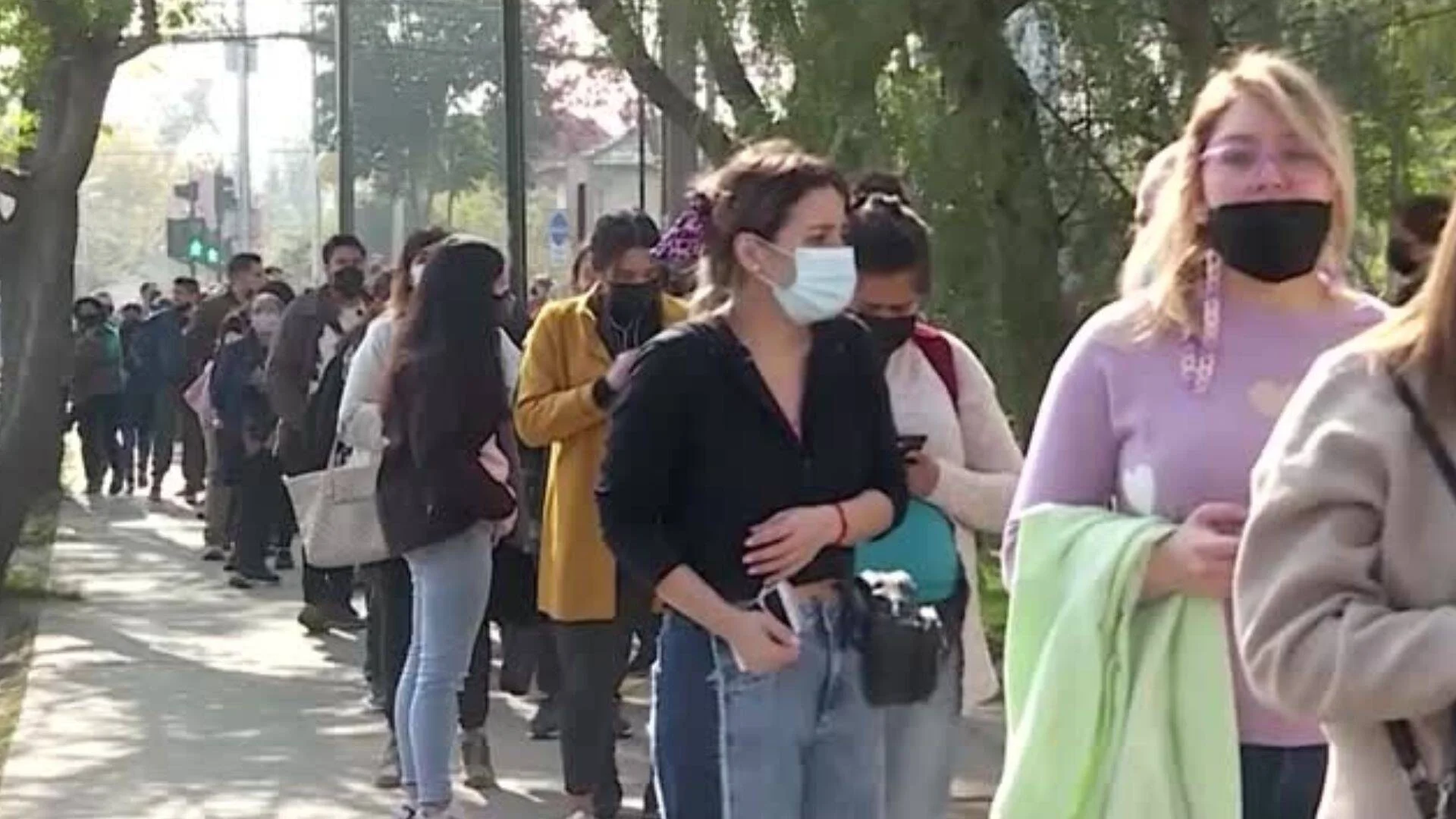 Numerosas personas con tapabocas hacen una larga fila sobre la vereda de una calle arbolada.