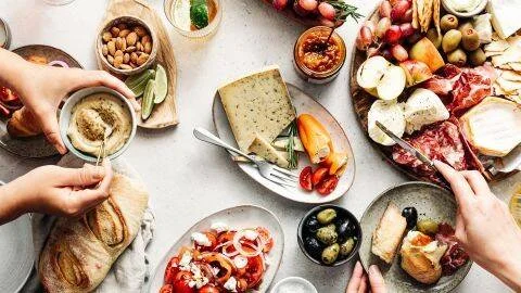 Manos sirviendo una mesa con tabla de quesos, fiambres, aceitunas, hummus, frutos secos y pan en un almuerzo compartido.