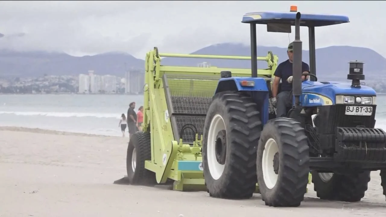 Un operario conduce un tractor New Holland con máquina limpiadora de arena en una playa, con la ciudad al fondo.