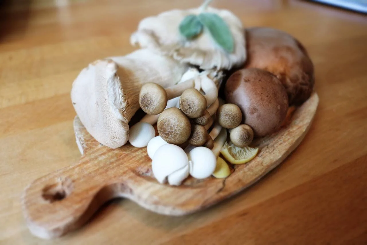 Distintas variedades de hongos frescos —blancos, shiitake y portobello— sobre una tabla de madera rústica con hojas de salvia.