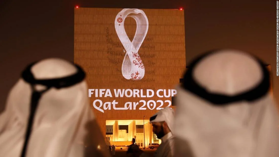 Dos hombres con vestimenta árabe tradicional observan el logo oficial de la FIFA World Cup Qatar 2022 proyectado en una fachada iluminada.