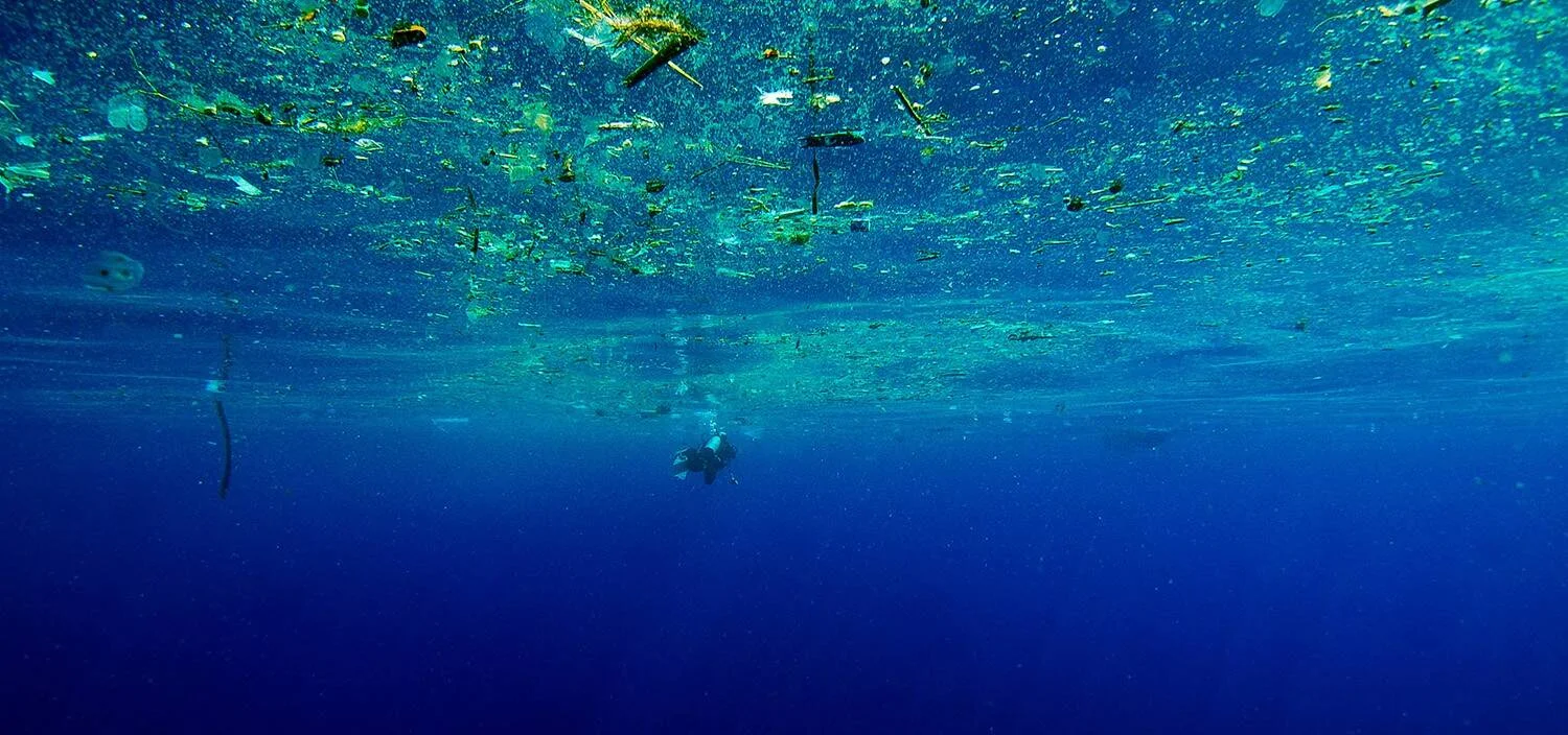 Vista submarina de un buzo rodeado de una densa capa de residuos plásticos flotando en la superficie del océano.