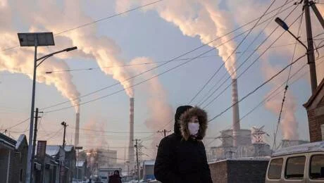 Persona con mascarilla y ropa de abrigo camina frente a múltiples chimeneas industriales emitiendo humo en día frío.