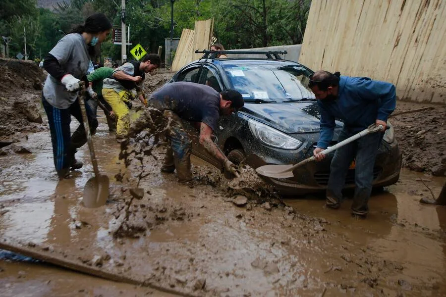 Varias personas usan palas para liberar un automóvil atrapado en el barro tras un aluvión en zona urbana.