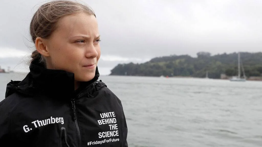 G. Thunberg con chaqueta negra que dice 'Unite Behind the Science #FridaysForFuture', mirando al horizonte frente al mar.