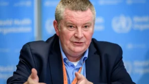 Hombre con credencial naranja de la OMS gesticula mientras habla frente a un fondo azul con el logo de la Organización Mundial de la Salud.