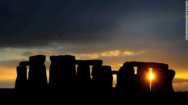 Silueta de Stonehenge recortada contra un cielo naranja al atardecer, con el sol asomando entre dos piedras verticales.