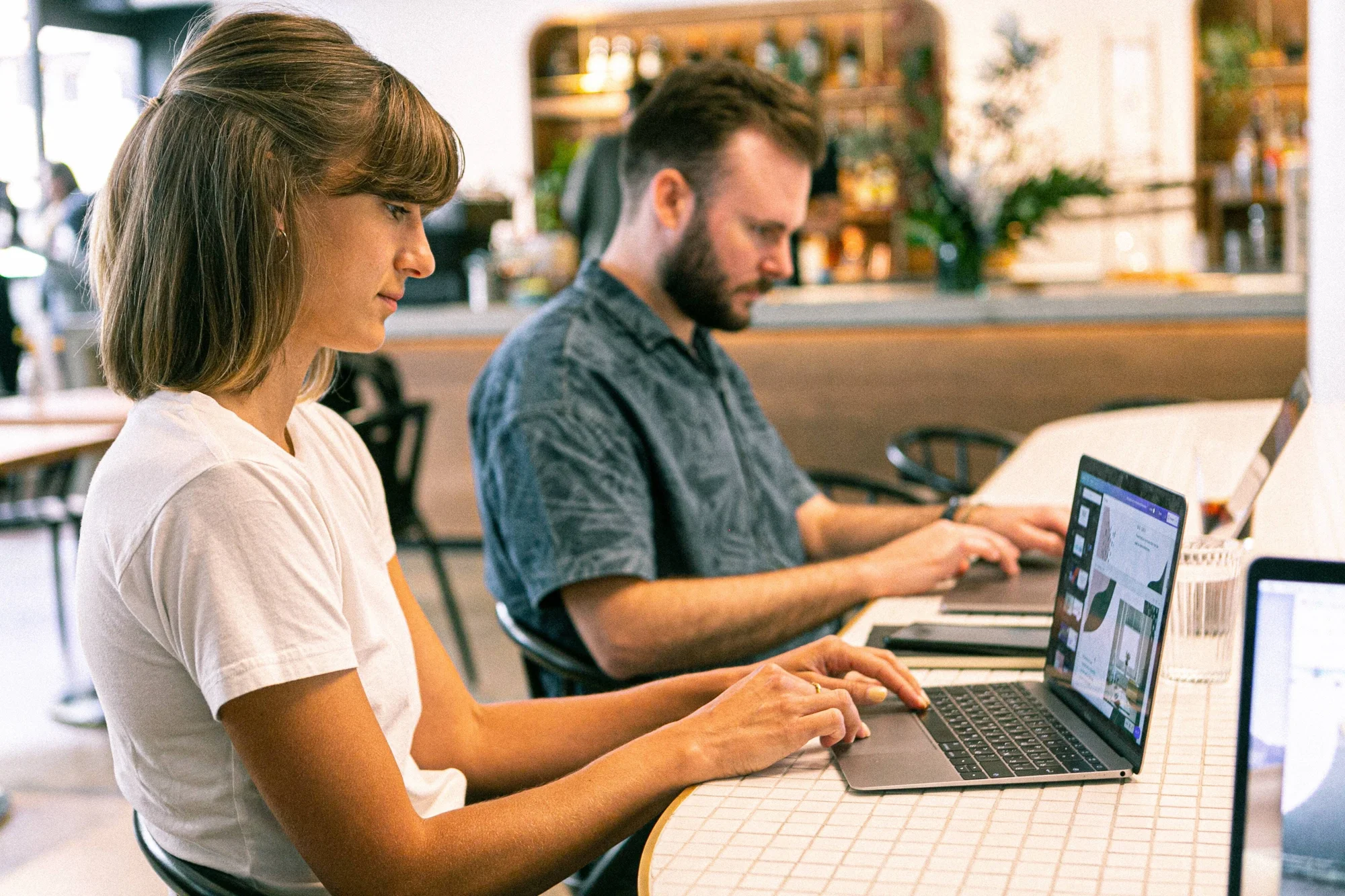 Una mujer y un hombre trabajan concentrados en sus laptops sentados en una mesa de cafetería moderna.