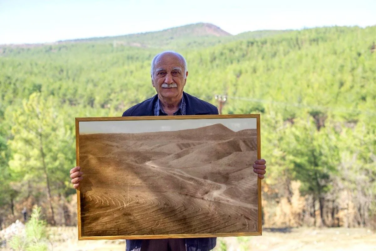 Hombre mayor muestra una fotografía enmarcada de colinas erosionadas y áridas, con un bosque verde frondoso de fondo, evidenciando el contraste antes y después de una reforestación.