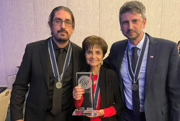 Tres personas con medallas al cuello posan junto a un trofeo de cristal en una ceremonia de premiación.