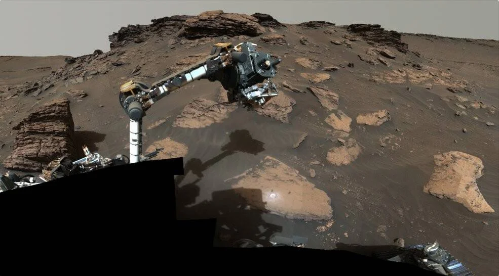 El brazo robótico de un rover extiende sus instrumentos sobre rocas y suelo rocoso de Marte.