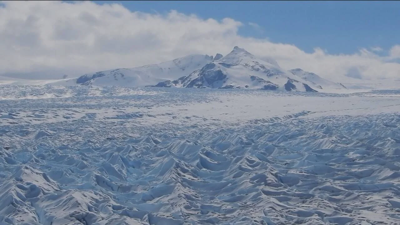 Vasta superficie glaciar con grietas de hielo azul y una montaña nevada bajo cielo parcialmente nublado.
