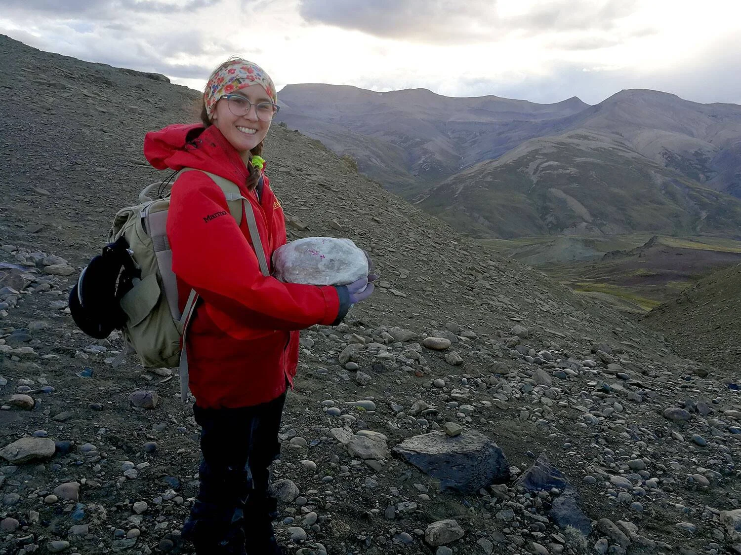 Investigadora con campera roja y mochila sonríe sosteniendo una muestra de roca marcada, en terreno pedregoso de alta montaña.