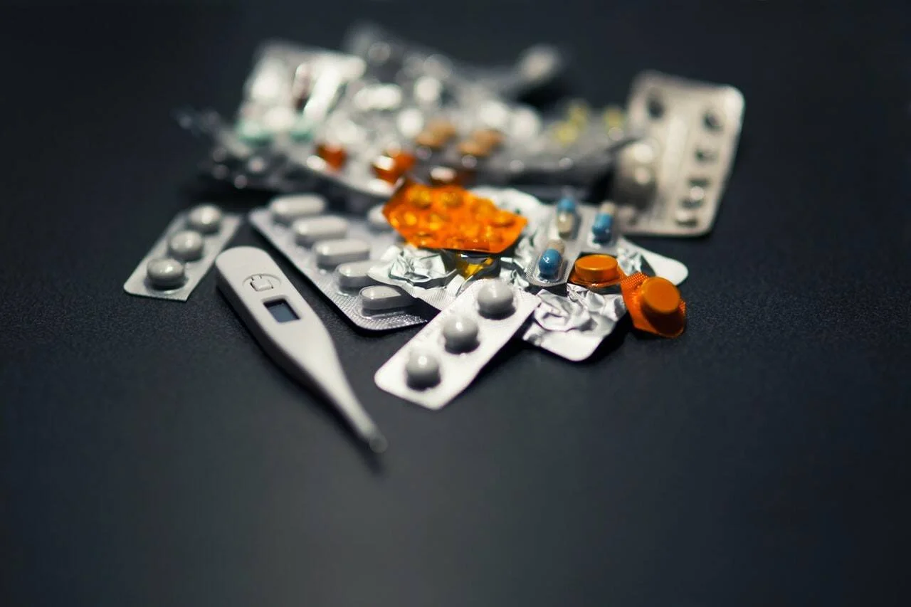 Varios blísteres de pastillas y cápsulas de colores junto a un termómetro digital sobre superficie oscura.