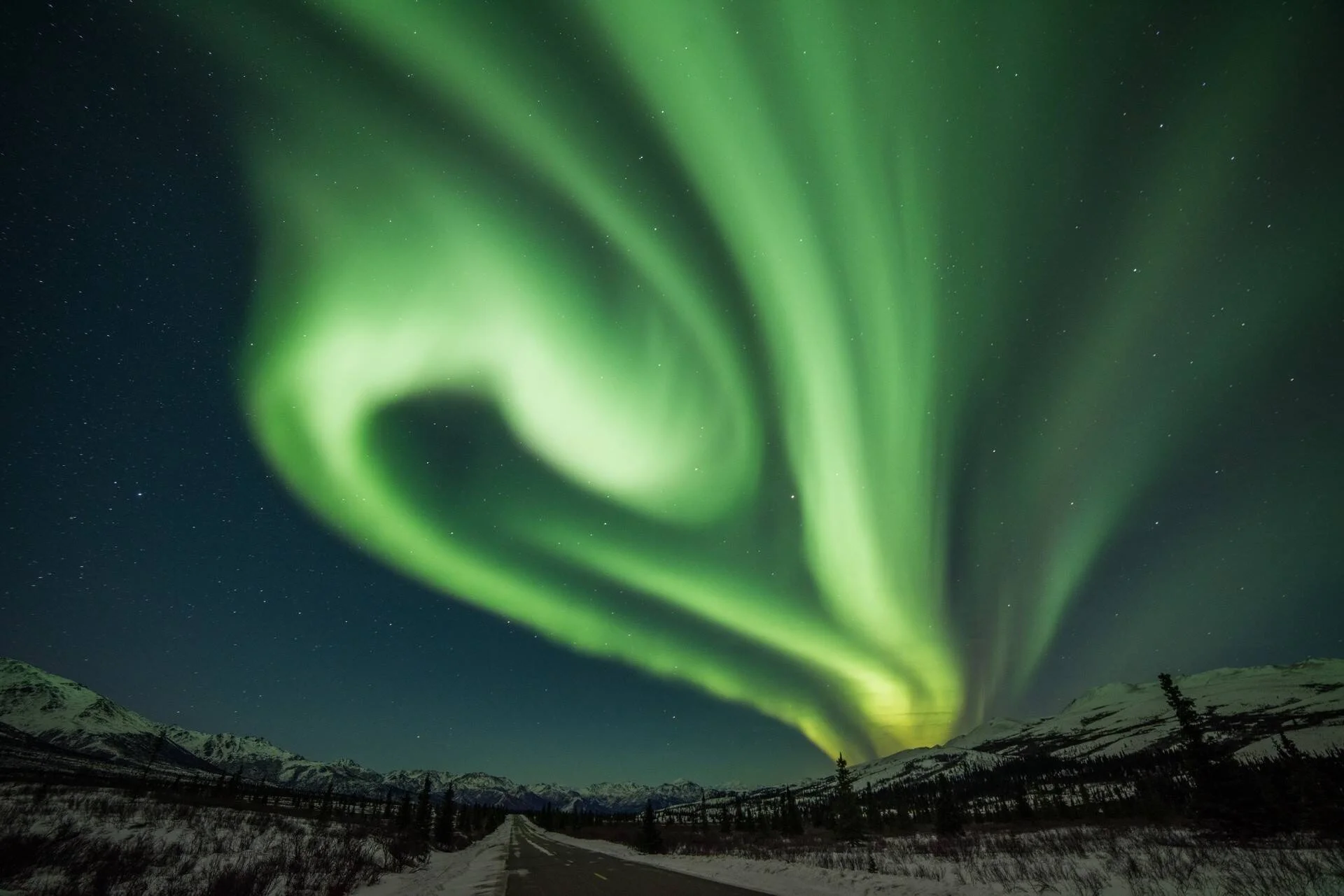 Espectacular aurora boreal verde en forma de espiral ilumina el cielo nocturno sobre una carretera nevada rodeada de montañas.