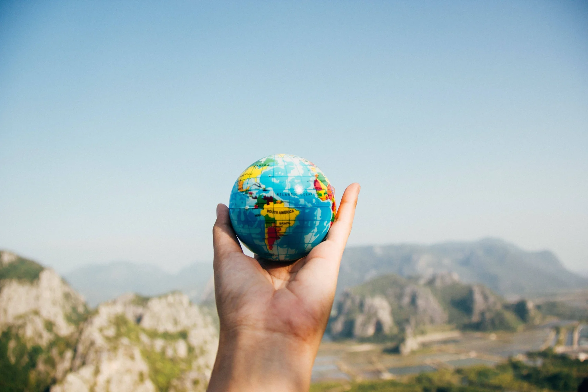 Una mano sostiene un pequeño globo terráqueo con un paisaje montañoso de fondo, simbolizando la conexión global.