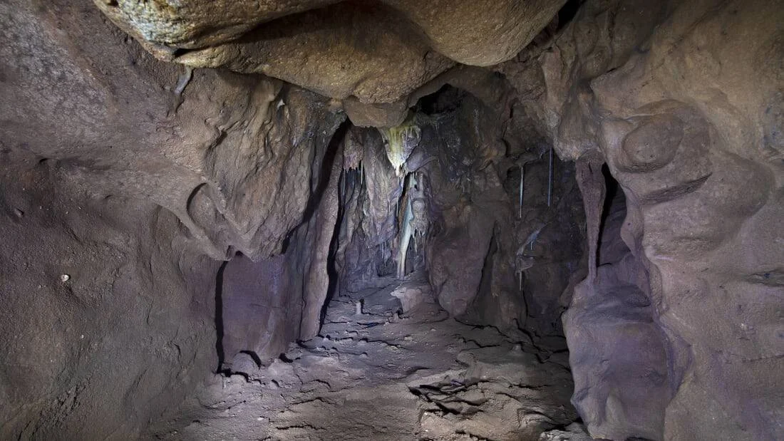 Pasaje interior de una cueva con paredes y techo de roca rojiza y estalactitas colgando del centro.