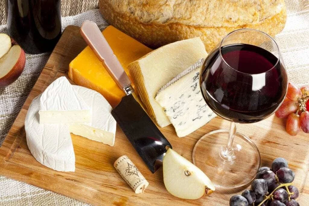 Tabla de madera con variedades de queso, una copa de vino tinto, uvas, pera, pan artesanal y un corcho.