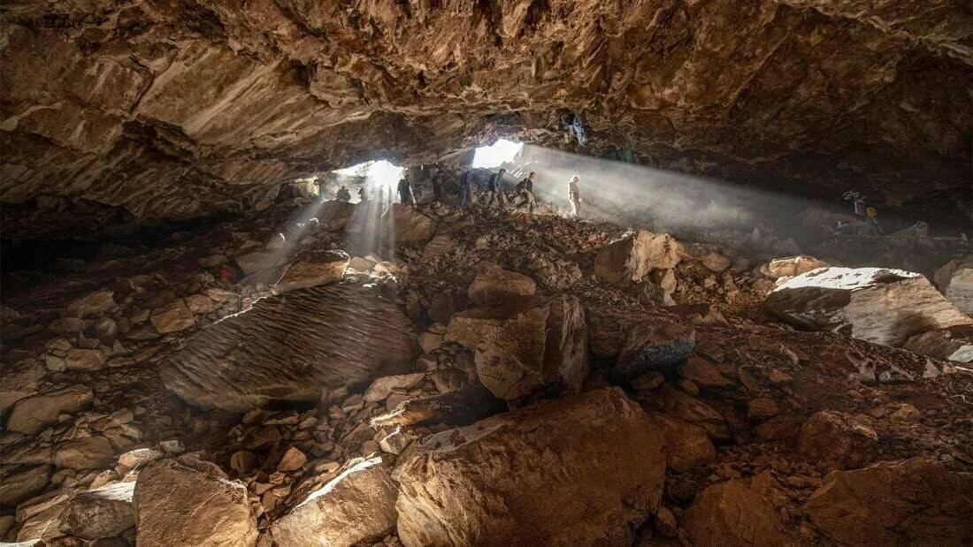 Rayos de luz solar penetran por la abertura de una gran cueva rocosa donde varias personas trabajan en su interior.