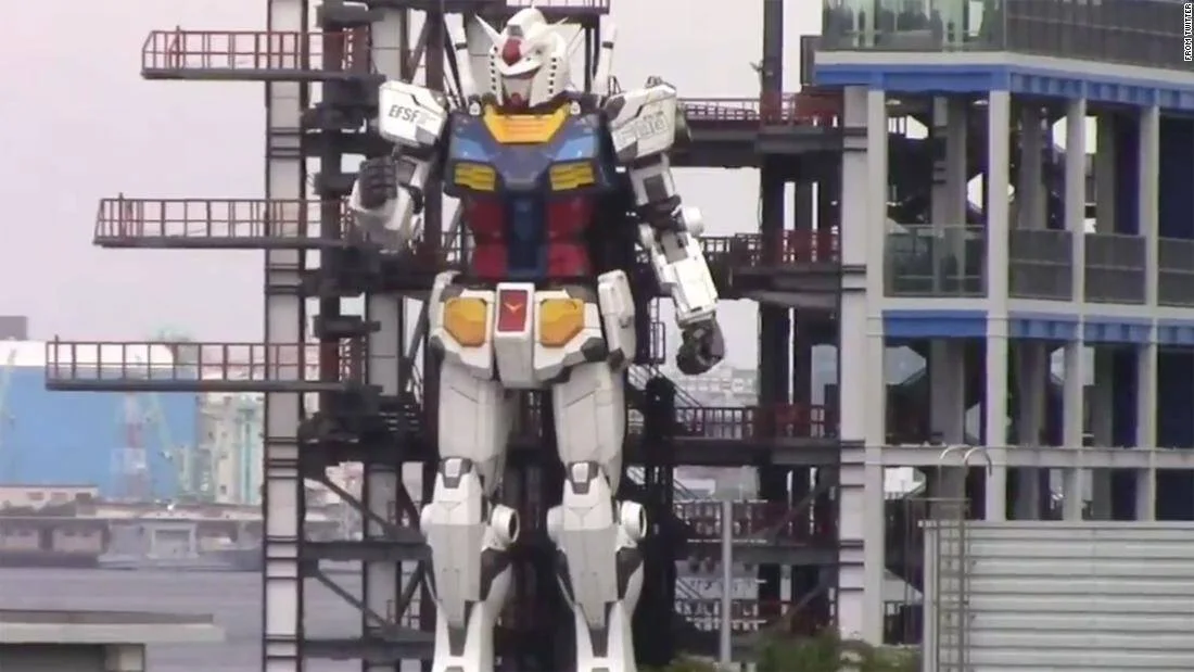 Enorme robot Gundam de tamaño real con armadura blanca, azul y amarilla, montado sobre estructura metálica en zona portuaria.