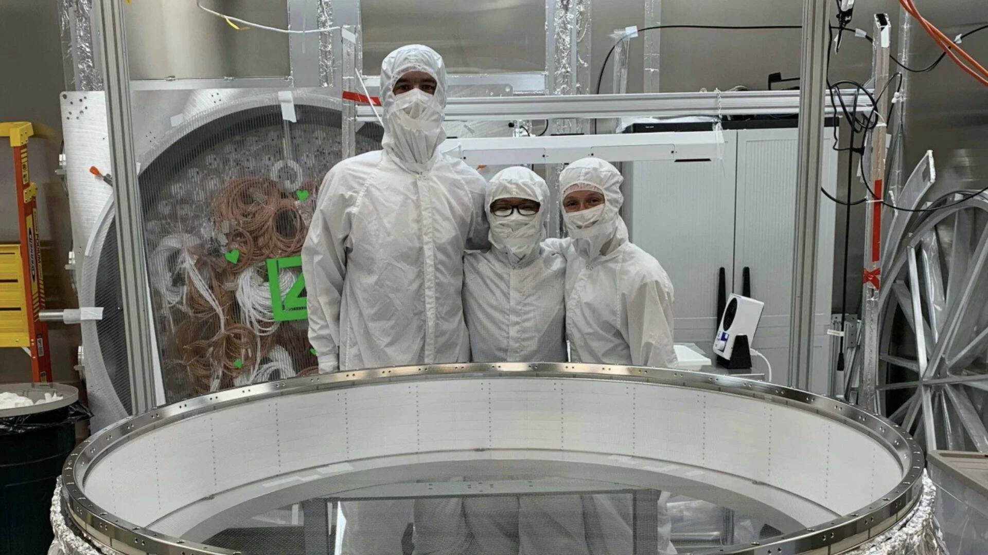 Tres personas con trajes de sala limpia posan junto a un gran componente circular metálico en un laboratorio de alta tecnología.