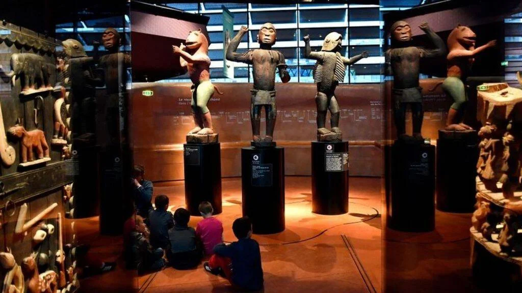 Grupo de niños sentados frente a estatuas africanas tradicionales expuestas en pedestales dentro de un museo de arte.