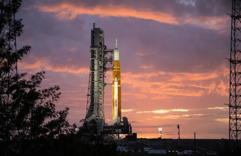 Cohete de la NASA iluminado en su plataforma de lanzamiento bajo un cielo rojizo al atardecer.