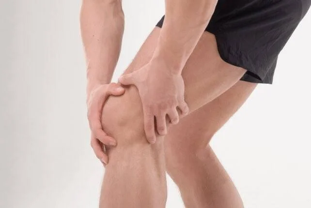 Hombre con ropa deportiva sostiene su rodilla con ambas manos, gesto que indica dolor o lesión en la articulación.