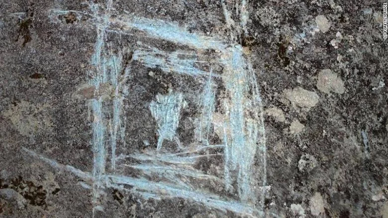 Rayados con tiza o pintura blanca azulada dañan un petroglifo ancestral grabado en roca oscura.