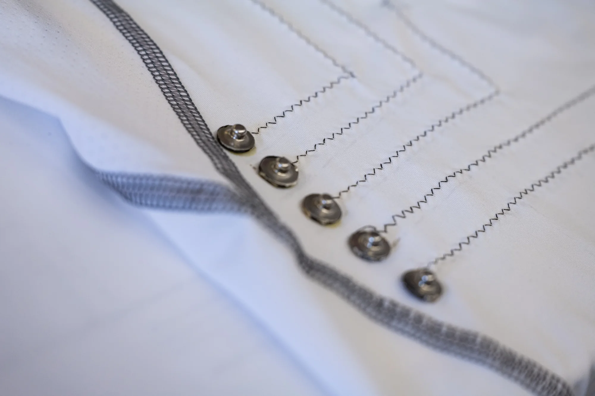Detalle de broches a presión metálicos sobre tela blanca con costuras en zigzag y cinta gris.