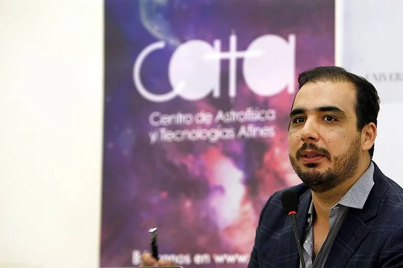 Un hombre con barba y saco habla frente al logo del Centro de Astrofísica y Tecnologías Afines (CATA).