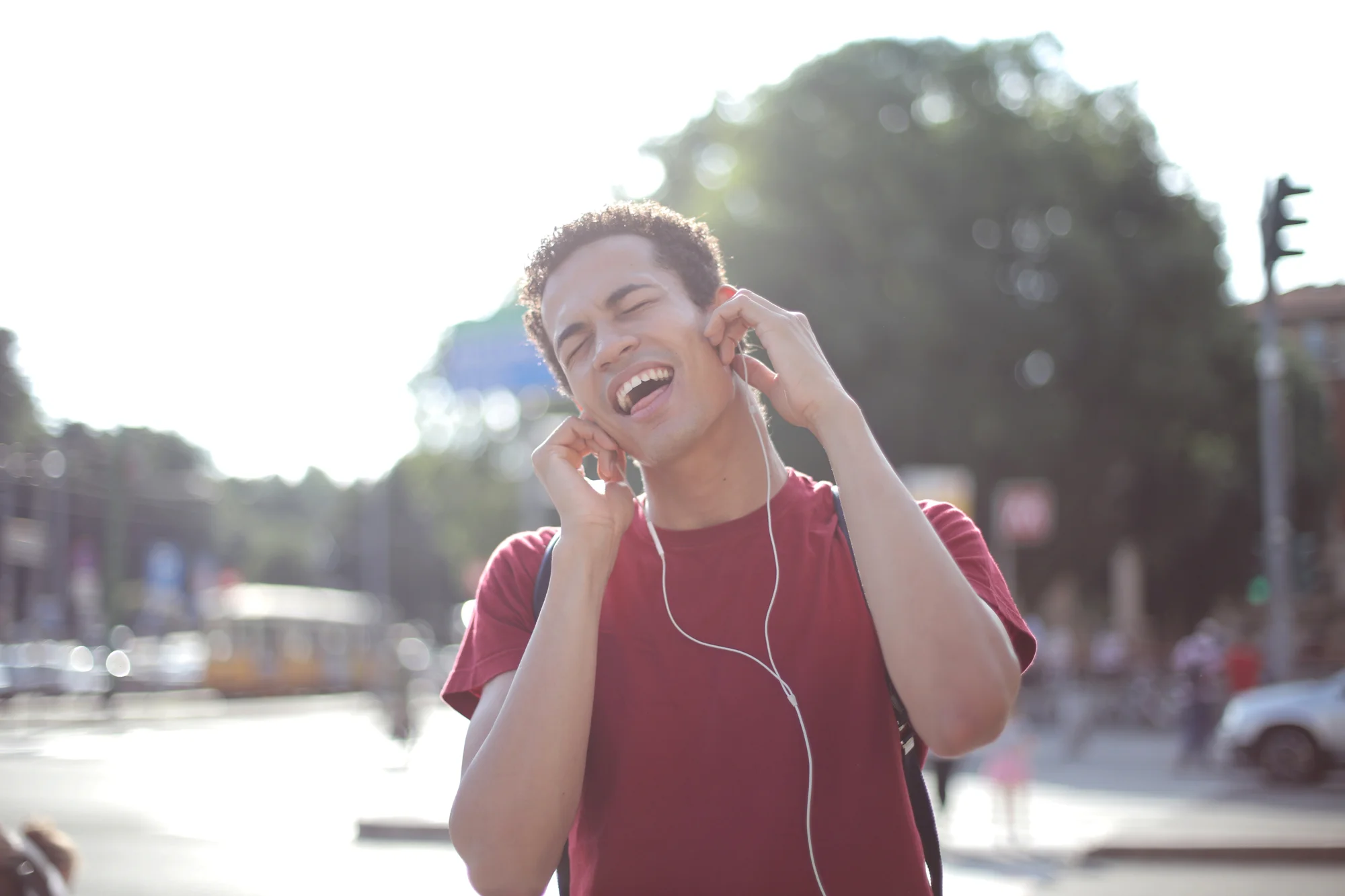 Joven sonriente con auriculares y mochila disfruta música en una calle urbana soleada.