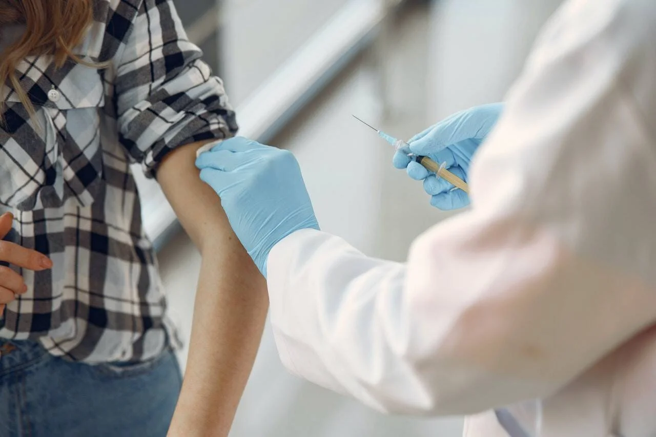 Un profesional sanitario con guantes azules aplica una inyección en el brazo de una paciente con camisa a cuadros.