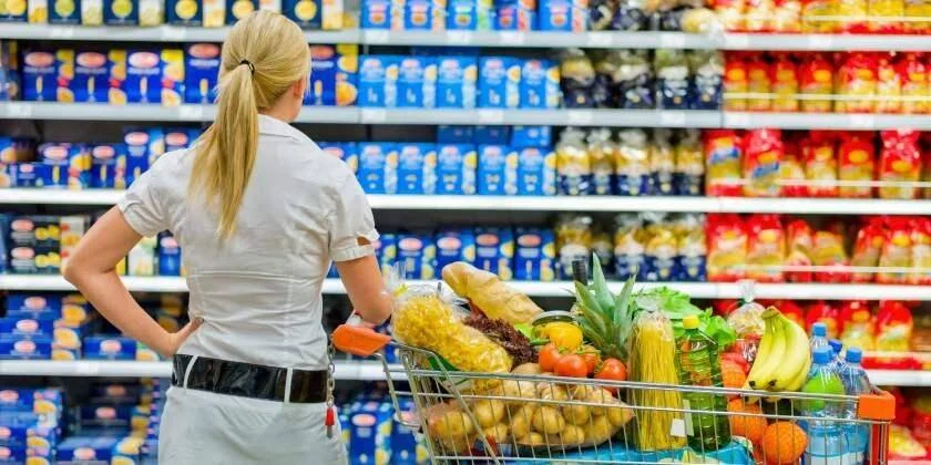 Mujer de espaldas observa góndolas de productos en un supermercado con su carrito cargado de frutas y alimentos.
