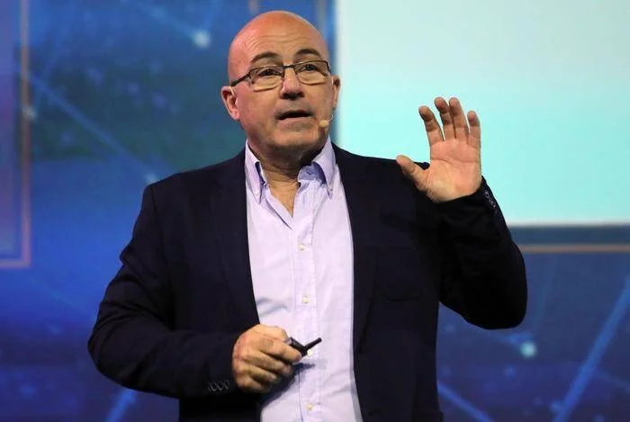 Hombre calvo con anteojos y saco azul habla en una conferencia con la mano levantada.