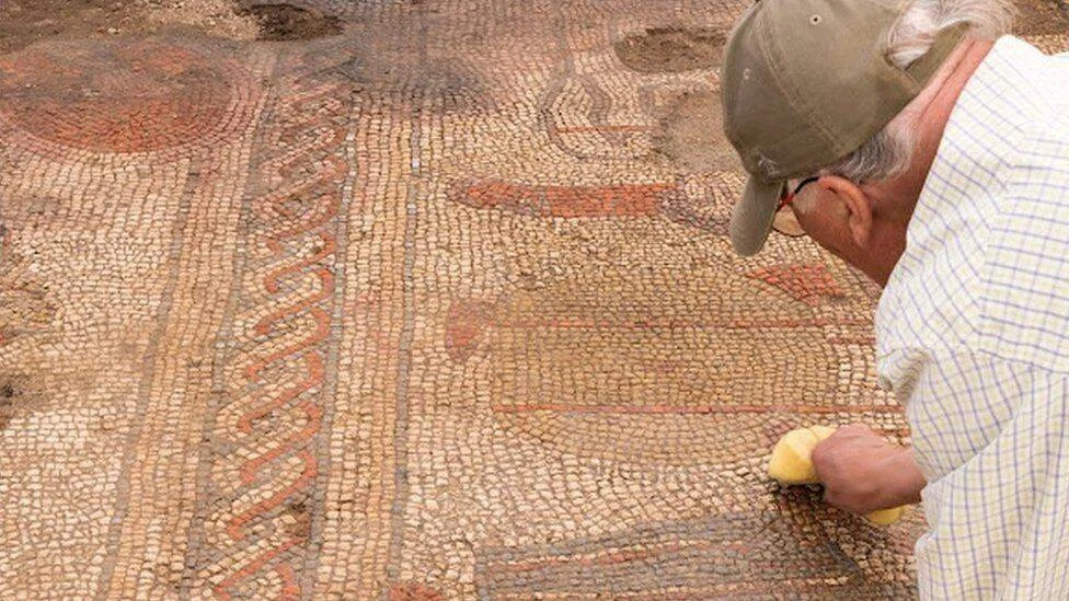 Un arqueólogo con gorra examina y limpia de cerca un mosaico romano con motivos geométricos en rojo y beige.