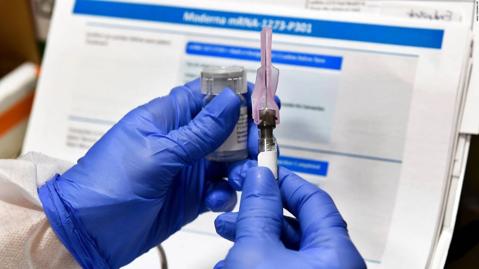 Manos con guantes azules extraen una dosis de un vial de la vacuna Moderna mRNA-1273 frente a un documento del ensayo clínico fase 3.