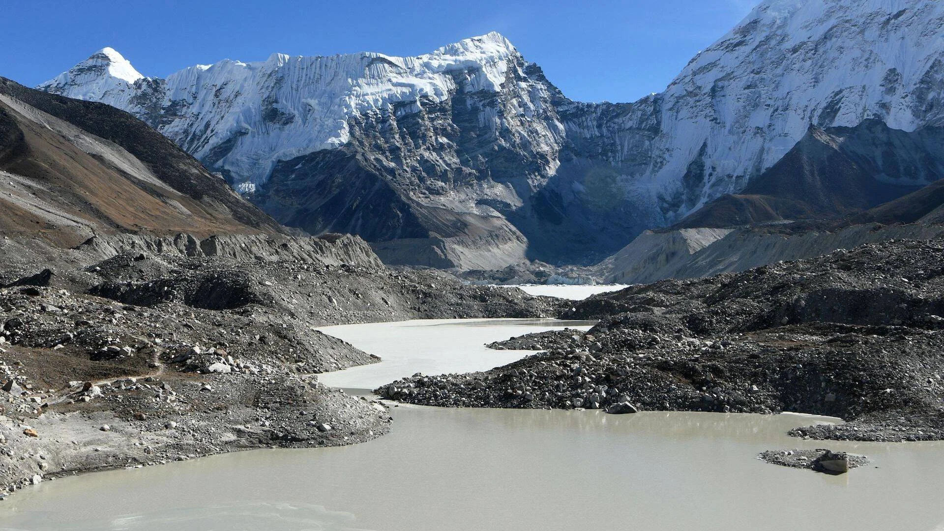 Lago de deshielo glaciar con agua turbia rodeado de morrenas rocosas y picos nevados del Himalaya al fondo.