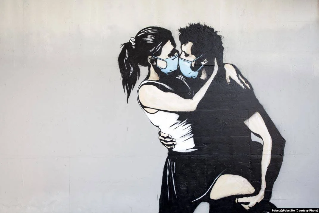 Grafiti en blanco y negro de una pareja abrazada y a punto de besarse, ambos con mascarillas quirúrgicas azules.