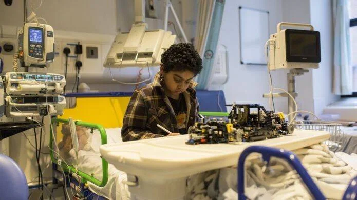 Adolescente armando una construcción de Lego junto a una cama de hospital rodeada de monitores médicos.
