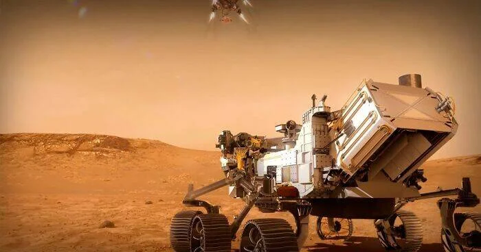 Rover espacial en la superficie rojiza de Marte con un pequeño helicóptero volando al fondo.