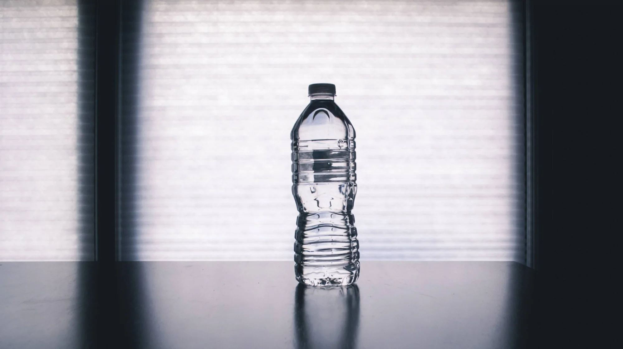 Botella de agua de plástico transparente con tapa negra sobre una mesa oscura frente a una persiana iluminada.