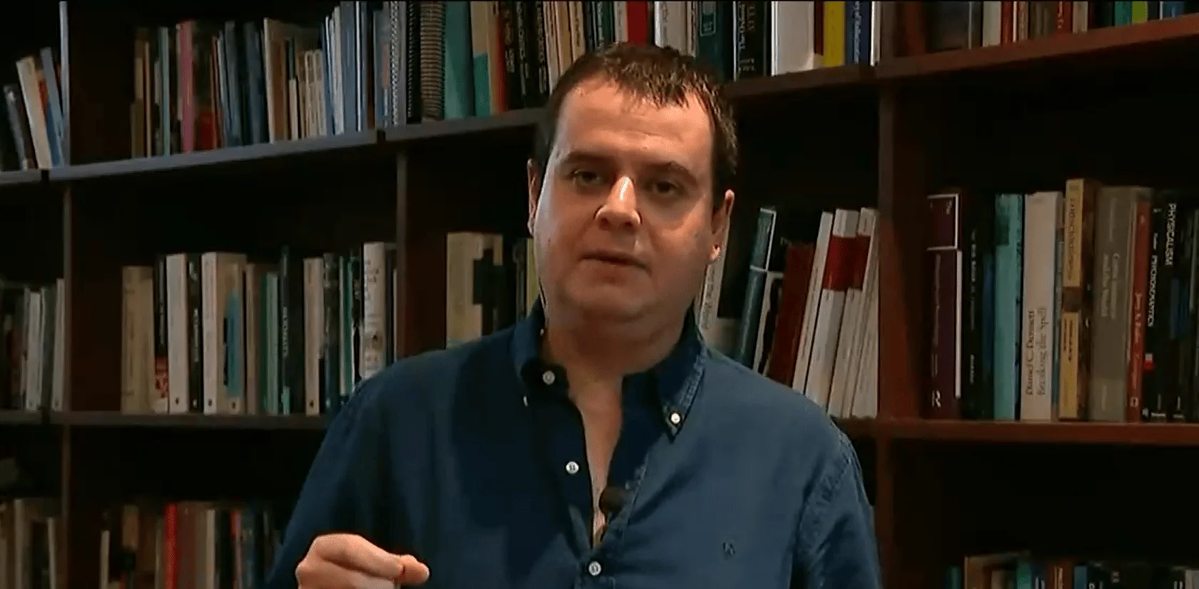 Hombre con camisa azul habla gesticulando frente a una gran estantería llena de libros.