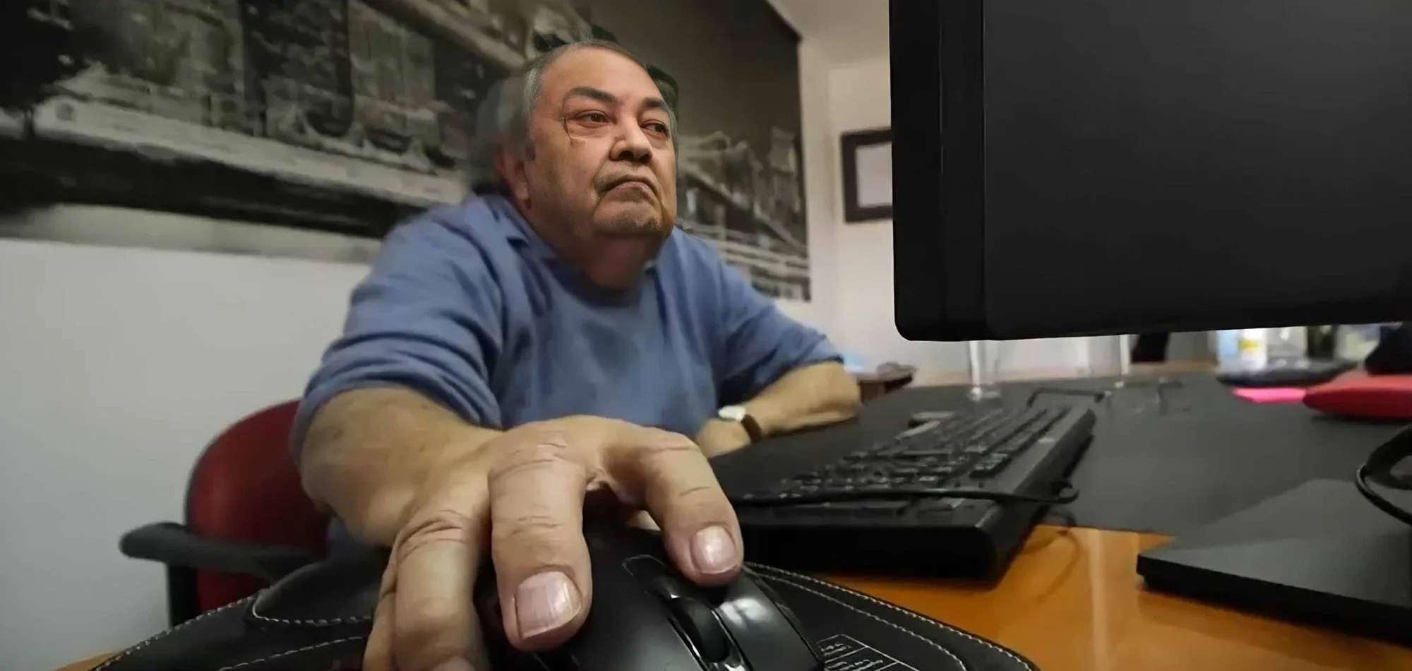 Hombre mayor con remera azul usando el mouse de una computadora en su escritorio de oficina.