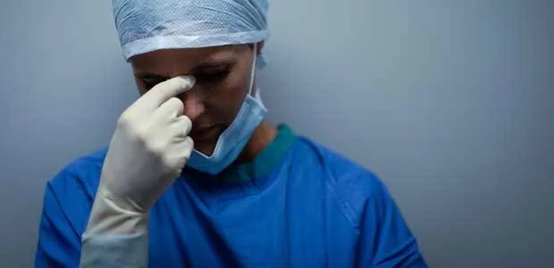 Profesional de la salud con uniforme quirúrgico azul y guantes se lleva la mano al rostro en gesto de agotamiento o angustia.
